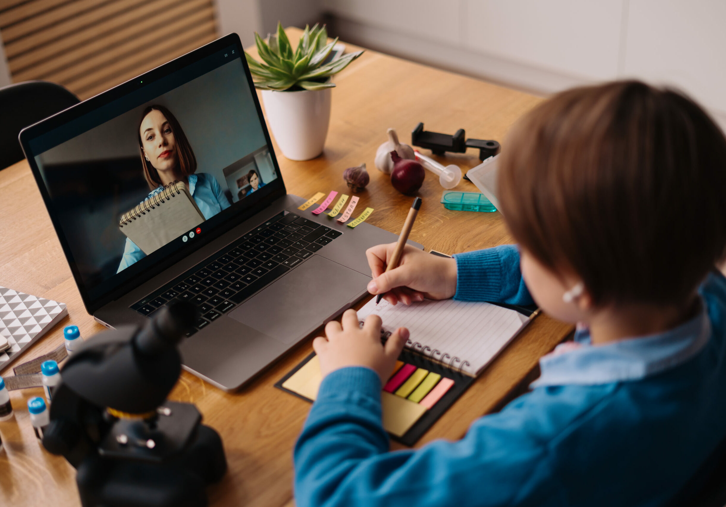 A Preteen boy uses a laptop to make a video call with his teacher. The Screen shows an online lecture with a teacher explaining the subject from class.